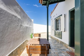 Un passadís exterior estret amb una banca de fusta i una taula, un sòl de rajola de pedra, i un edifici blanc amb una porta i persianes verdes.