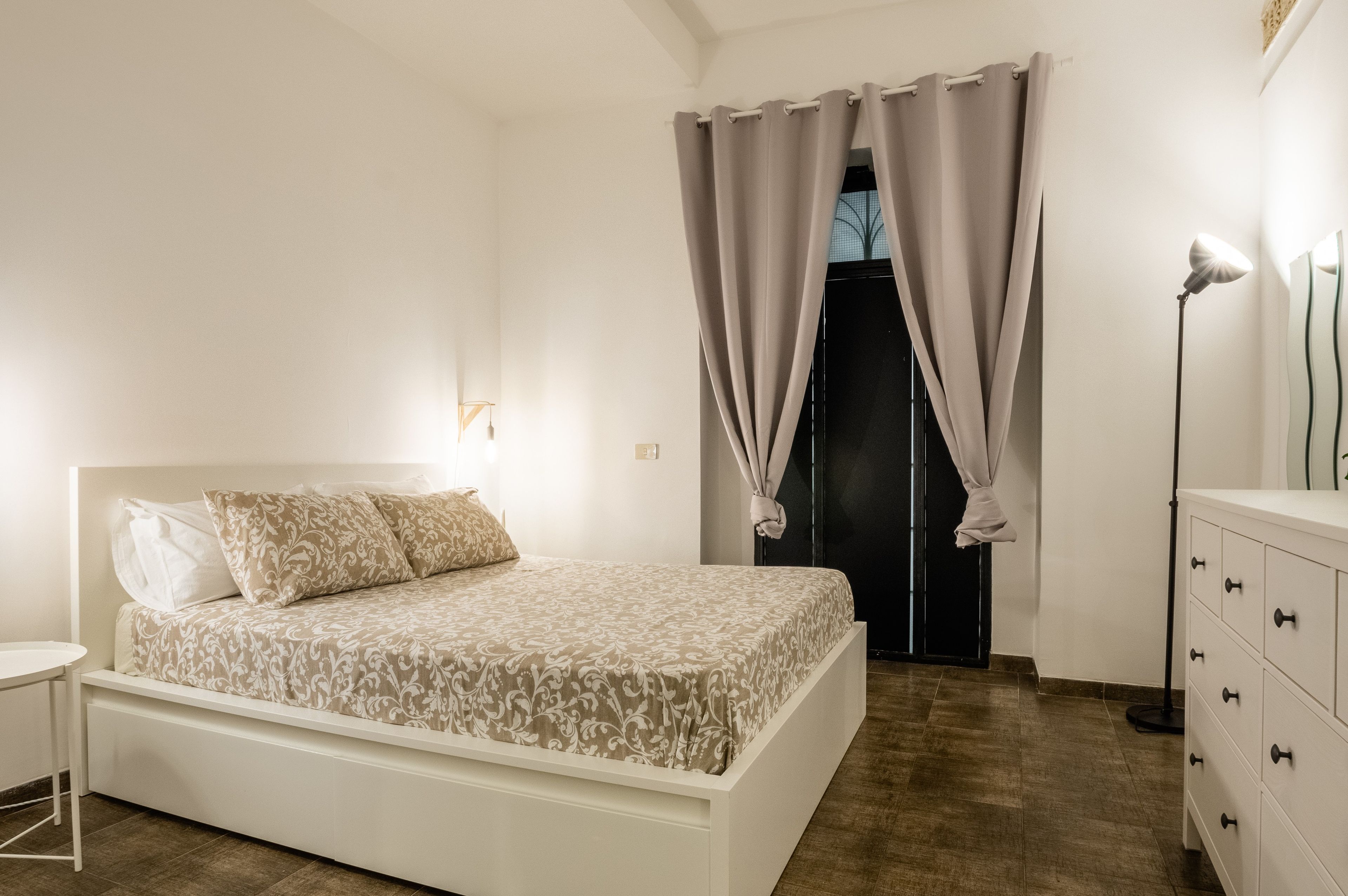 Une chambre à coucher avec un lit, une table de chevet, une commode et une porte avec des rideaux.