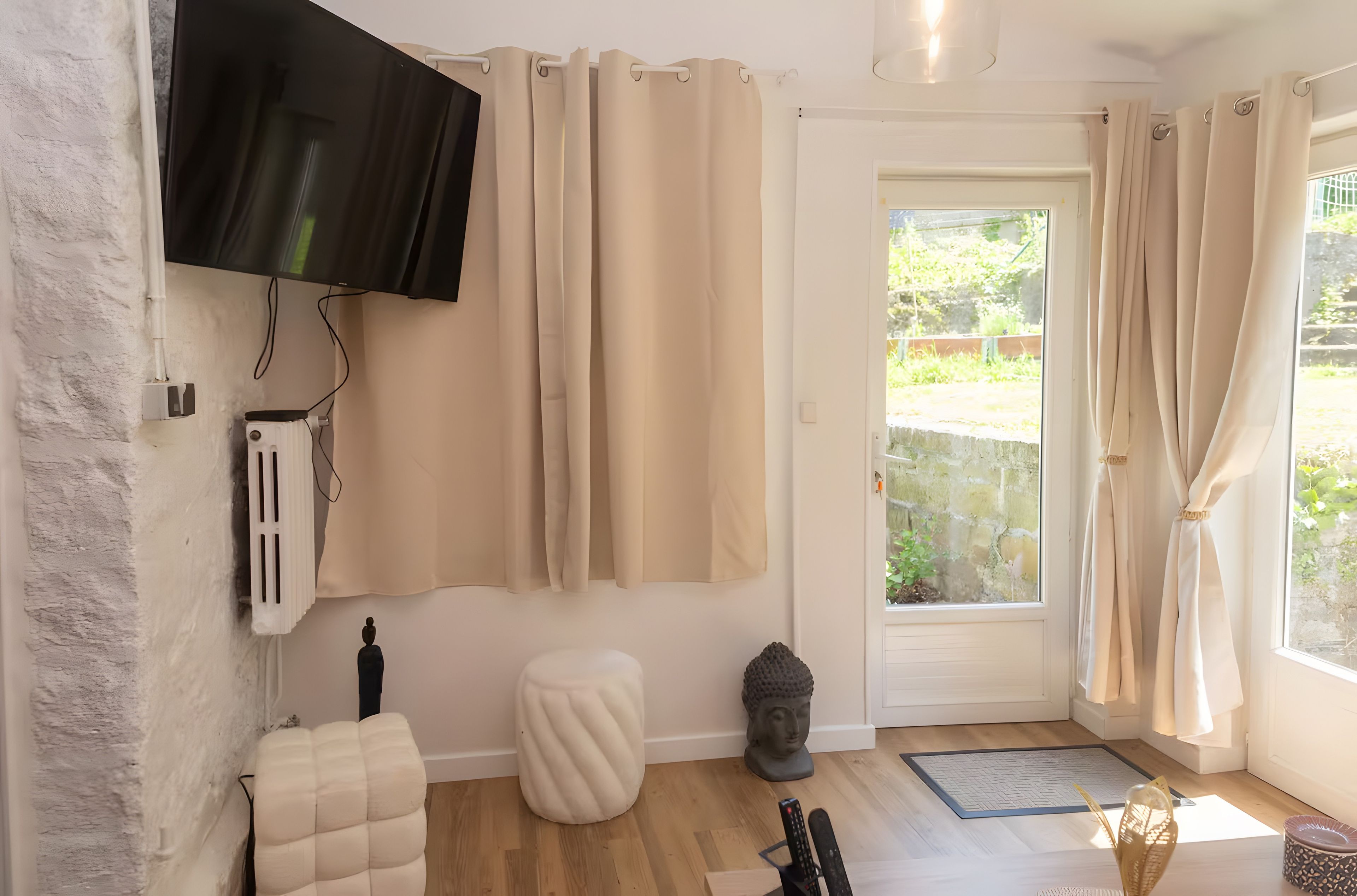 Een kamer met een flatscreen televisie aan de muur, beige gordijnen, een witte deur naar buiten, een klein wit krukje, een Boeddhabeeld en een afstandsbediening op de vloer.