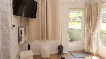 Een kamer met een flatscreen televisie aan de muur, beige gordijnen, een witte deur naar buiten, een klein wit krukje, een Boeddhabeeld en een afstandsbediening op de vloer.