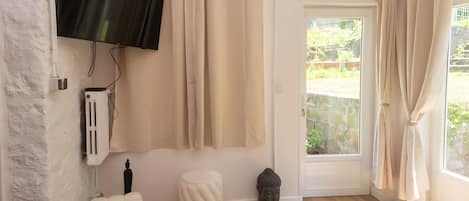 A room with a flat-screen TV mounted on the wall, beige curtains, a white door leading outside, a small white stool, a Buddha statue, and a remote control on the floor.