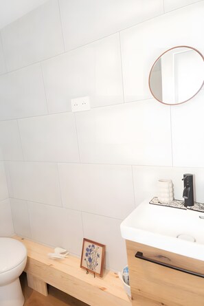 Un baño con pared de azulejos blancos, un espejo redondo, un lavabo blanco con grifo negro y un banco de madera con un cuadro enmarcado.