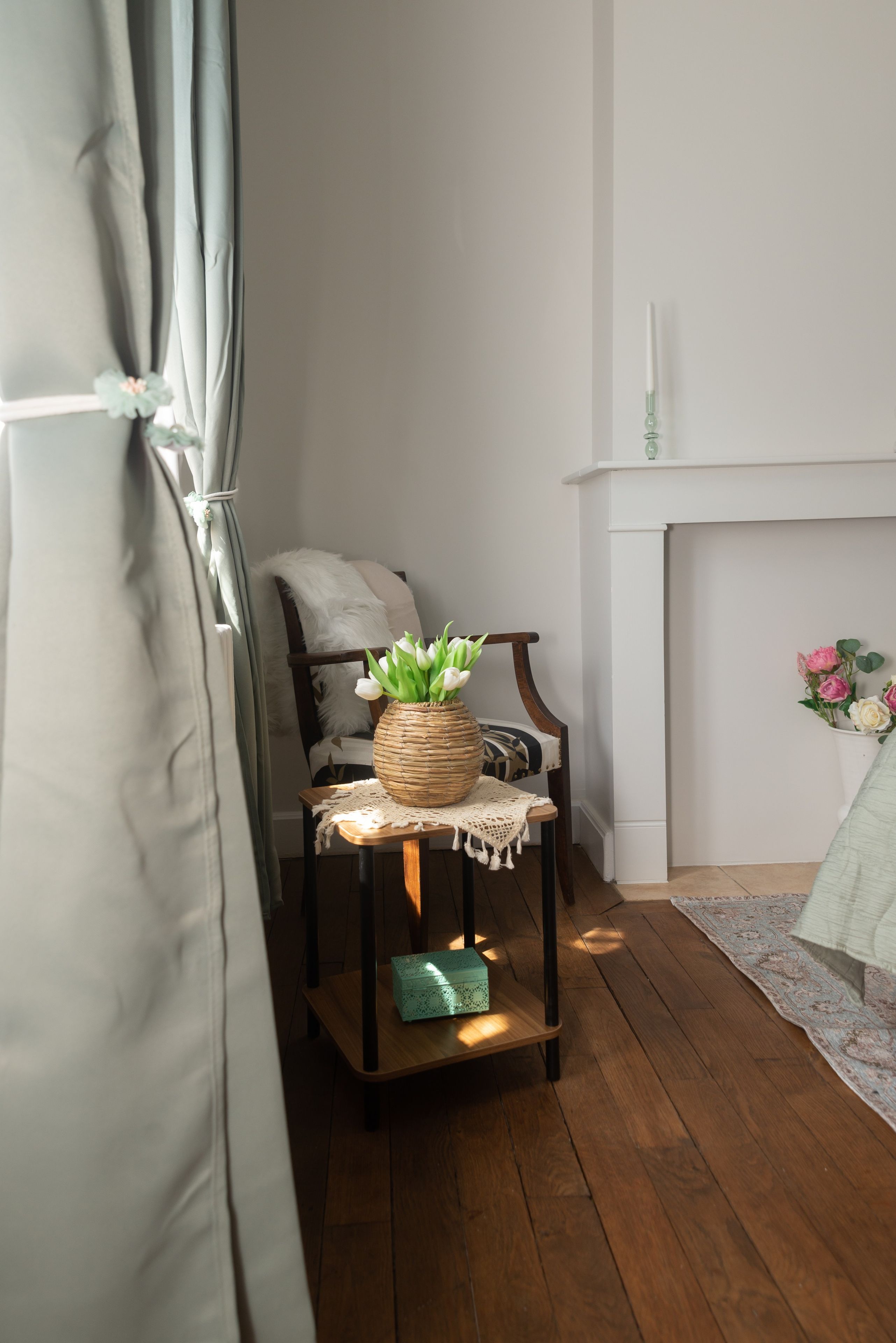 A room with a wooden floor, a chair with a white cushion, a wicker table with a vase of flowers, and a fireplace with a white mantel.