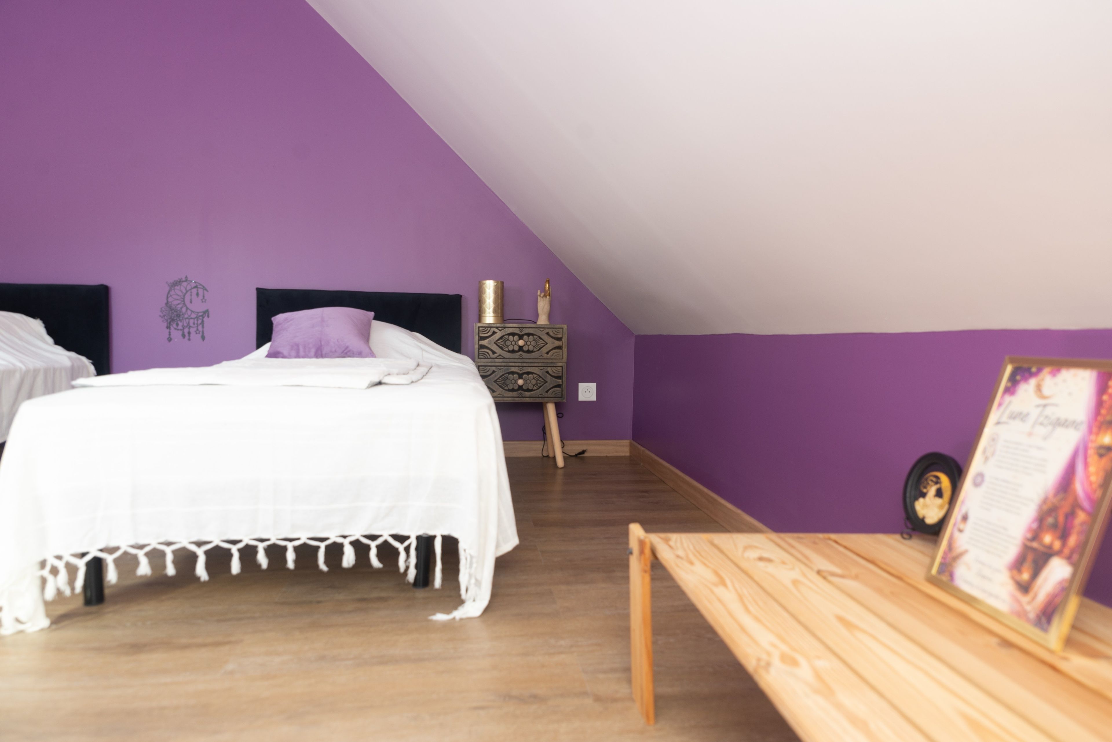 Een slaapkamer met een schuine kap, een bed met witte beddengoed, een houten bankje en een wand met een decoratieve dreamcatcher.