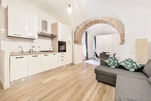 Una cucina moderna con mobili bianchi, piano cottura a gas e forno. C'è un'area pranzo con tavolo e sedie, e un soggiorno con divano e cuscini decorativi.