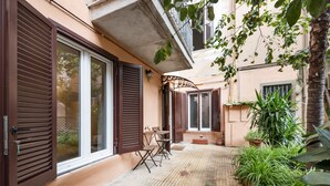 Un patio con suelo de baldosas, una mesa de madera y sillas, rodeado de plantas en macetas y una palmera.