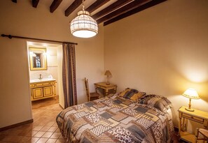 Une chambre à coucher avec un grand lit, une table de chevet avec une lampe, une tête de lit en bois, une fenêtre avec des rideaux et une salle de bain avec un lavabo et un miroir.