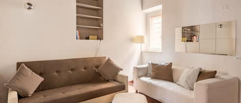 Un salon avec un canapé marron, un canapé blanc et une petite table sur laquelle se trouve une lampe. Il y a une fenêtre avec des stores et un miroir accroché au mur.