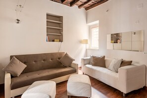 Ein Wohnzimmer mit braunem Sofa, weißem Sofa und einem kleinen Tisch mit einer Lampe. Ein Fenster mit Jalousien und ein Spiegel an der Wand sind ebenfalls vorhanden.