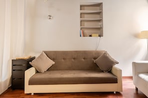 Un canapé aux couleurs neutres, agrémenté de deux coussins, une petite table d’appoint et une étagère murale avec des livres et une lampe.