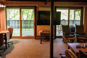 A room with a wooden ceiling, a carpeted floor, a table with chairs, a television, and a view of a wooden deck through large glass doors.