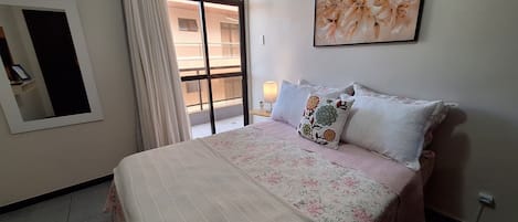 Une chambre à coucher avec un lit, une fenêtre avec des rideaux, une lampe fixée au mur et une œuvre d’art florale encadrée.