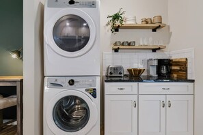 A compact kitchen with a stacked washer and dryer, a toaster, a coffee maker, and wooden shelves with kitchenware. - Shepard`s Lodge - families and pets welcome! (Indianapolis)