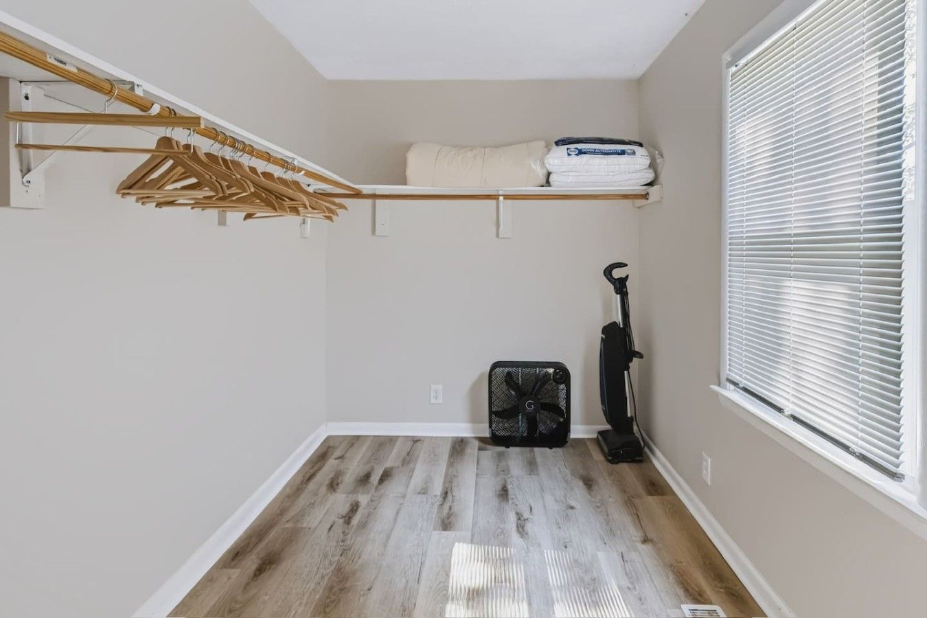 Une pièce avec un plancher en bois, un ventilateur de plafond, un aspirateur, une fenêtre avec des stores et un porte-vêtements avec des cintres.