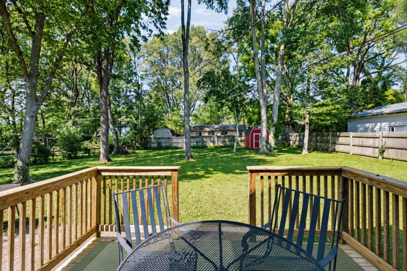 A deck with a metal table and chairs, surrounded by a wooden railing, overlooking a grassy yard with trees and a red shed.