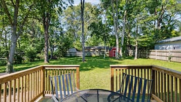 Une terrasse dotée d’une table et de chaises en métal, entourée d’une rambarde en bois, donnant sur une cour verdoyante avec des arbres et un abri de jardin rouge.
