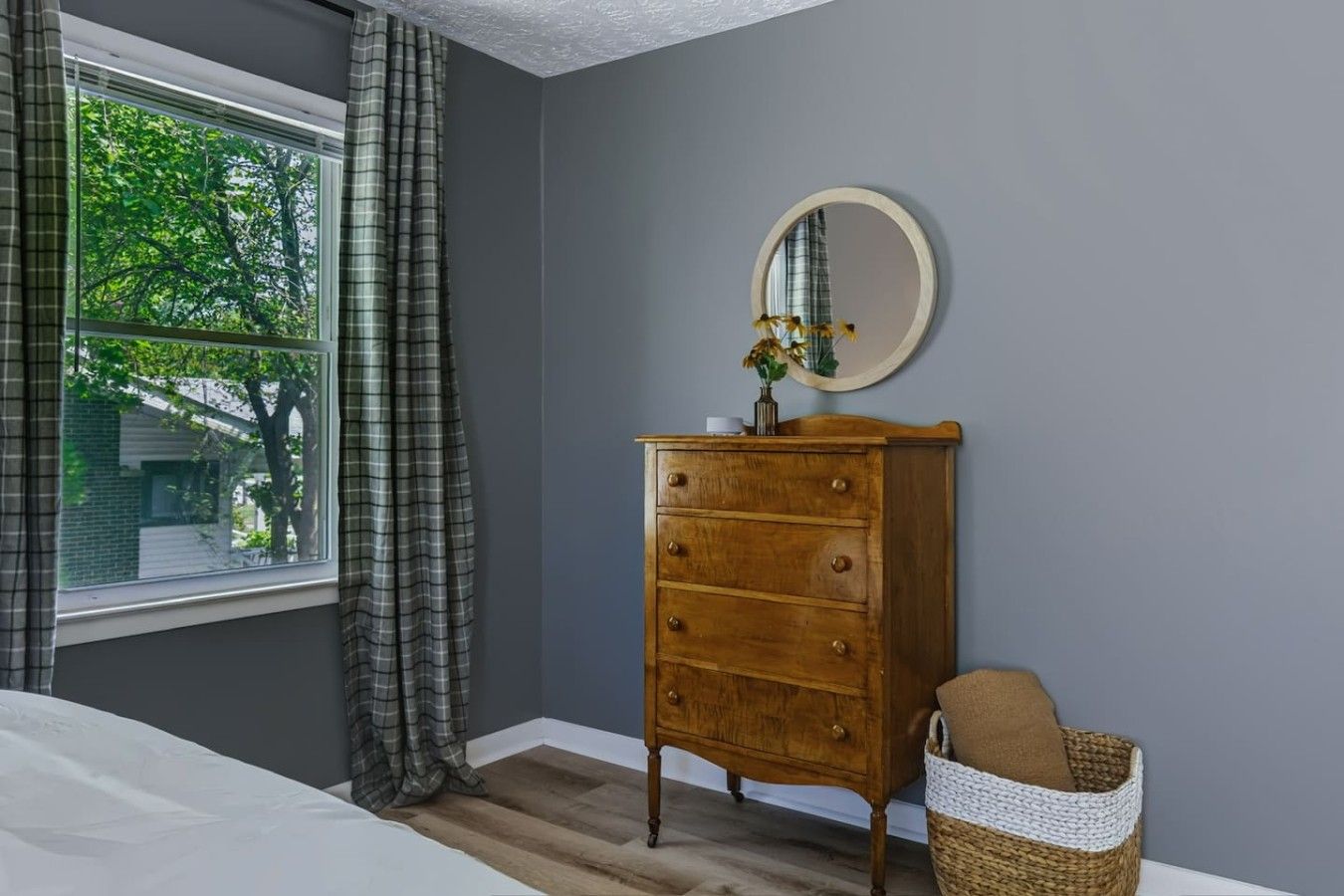 Quarto com cômoda de madeira, espelho, janela com cortinas e cesta de vime.