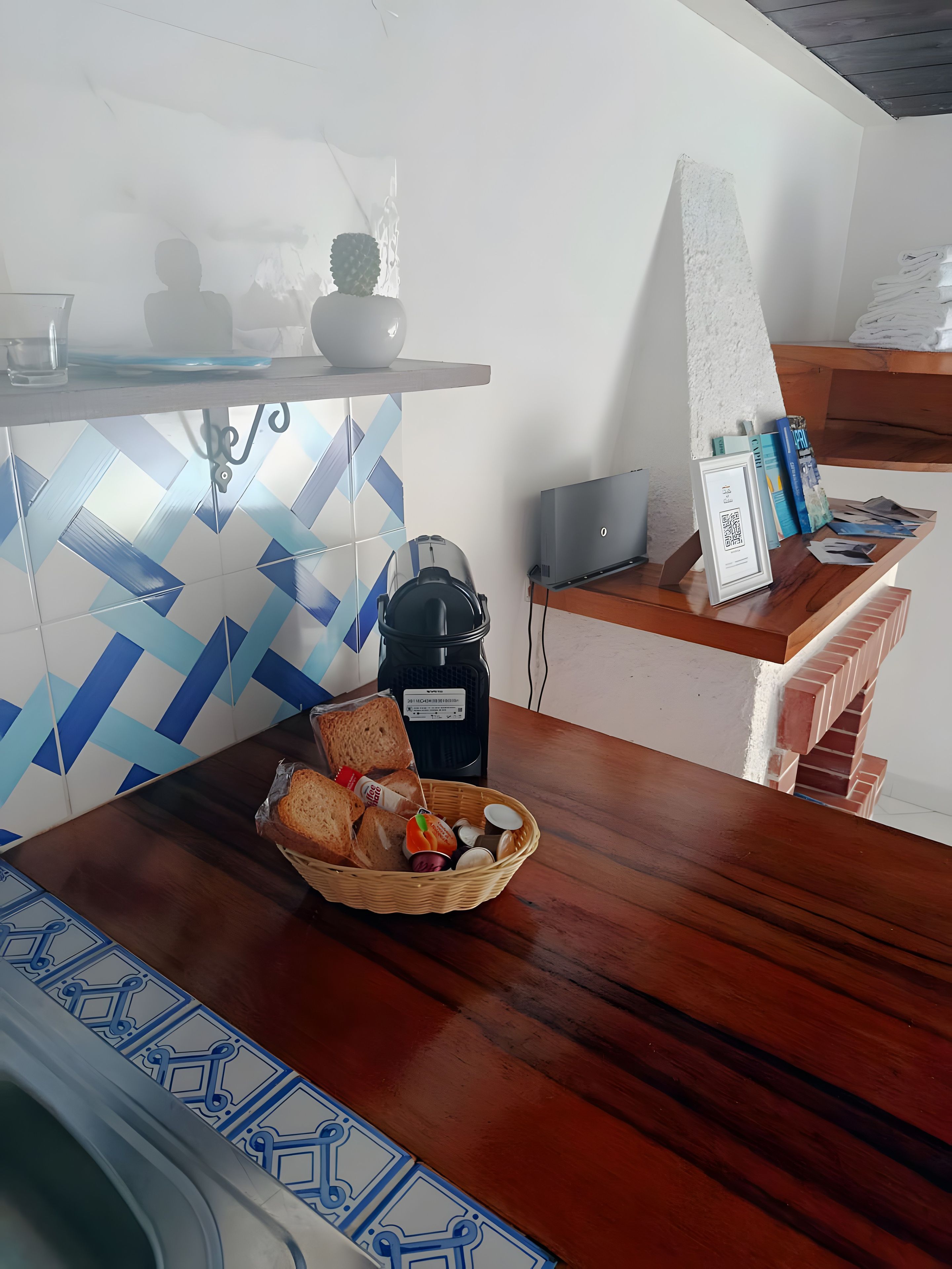 Una mesa de madera con una cesta de pan y condimentos, una cafetera y un ordenador portátil sobre una estantería.
