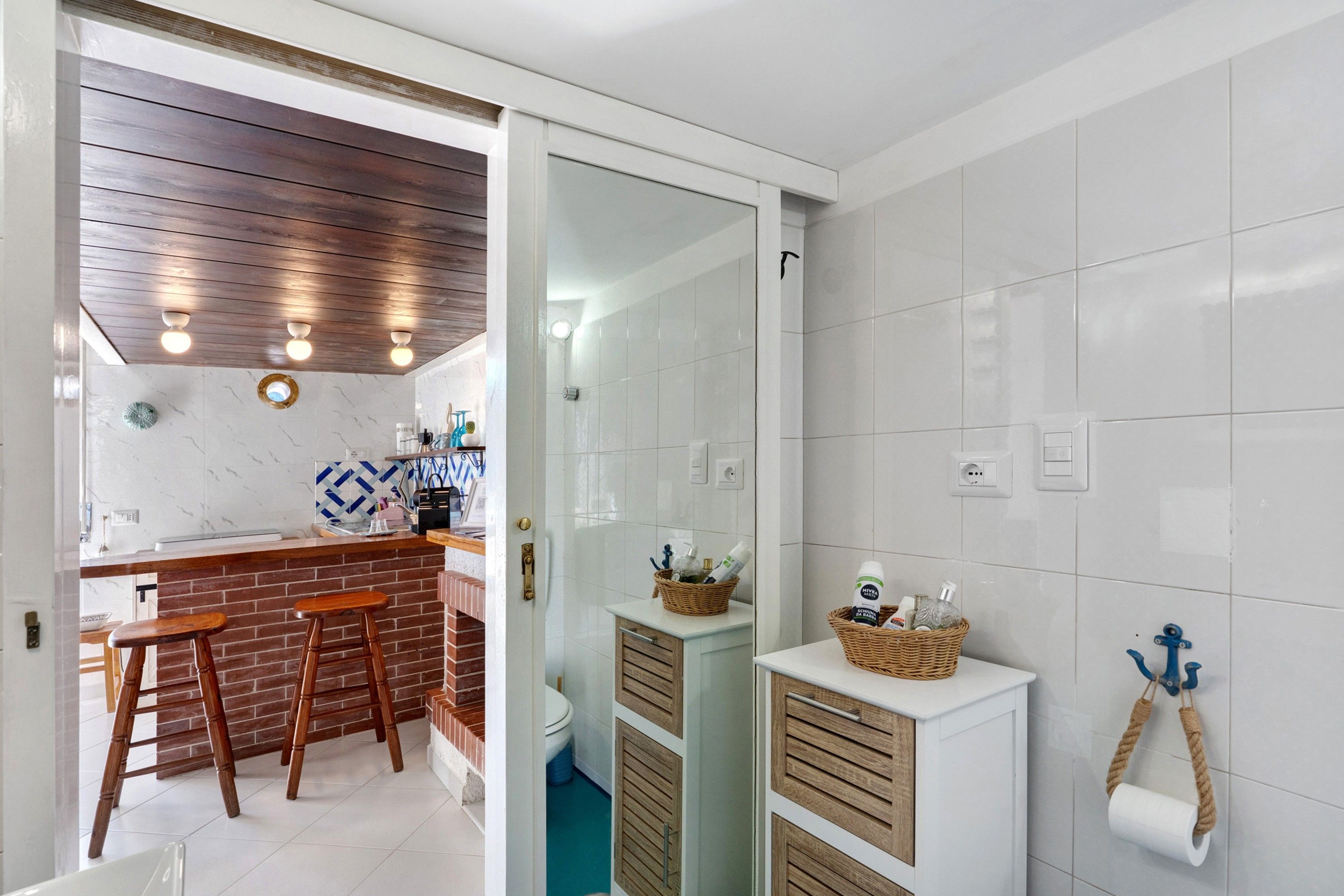 A modern bathroom with white tiles, a wooden ceiling, and a small sink area with stools.