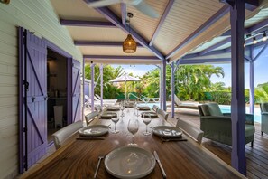 Un espace extérieur abrité, aménagé pour les repas, avec une table en bois dressée pour un repas, entourée de chaises, et donnant sur une piscine.