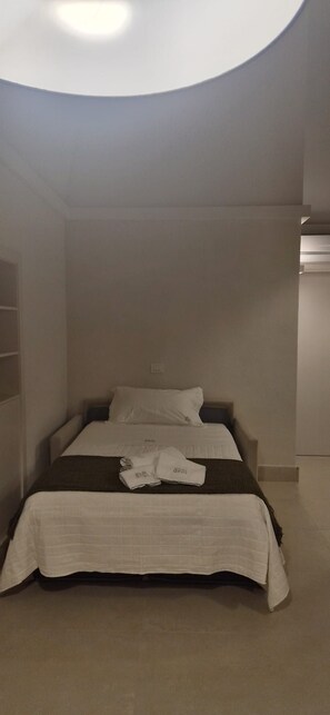 A single bed with a white bedspread and pillows, a dark throw blanket, and neatly folded towels on the bed.
