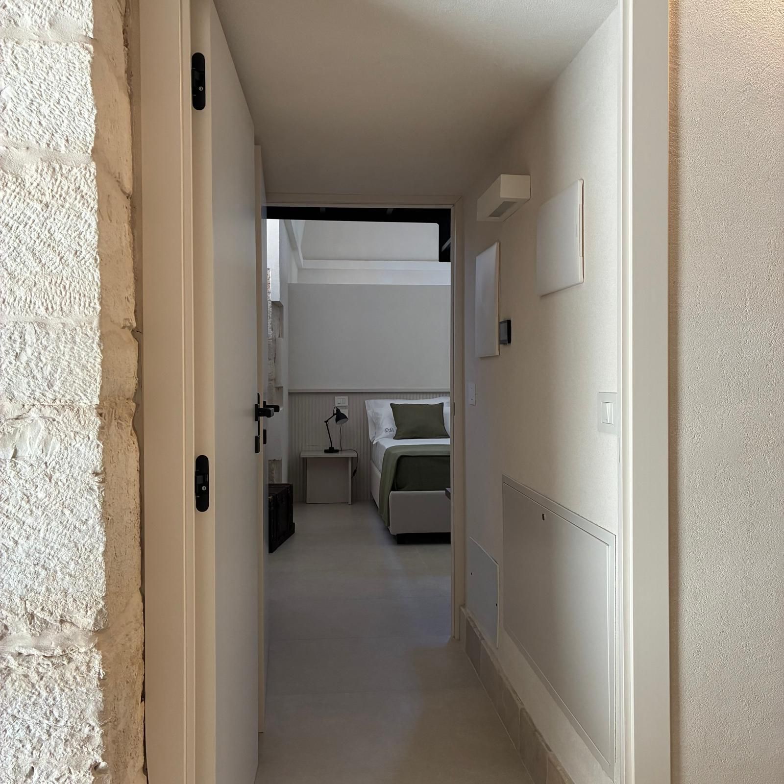 A narrow hallway leading to a bedroom with a bed, bedside table, lamp, and a radiator.