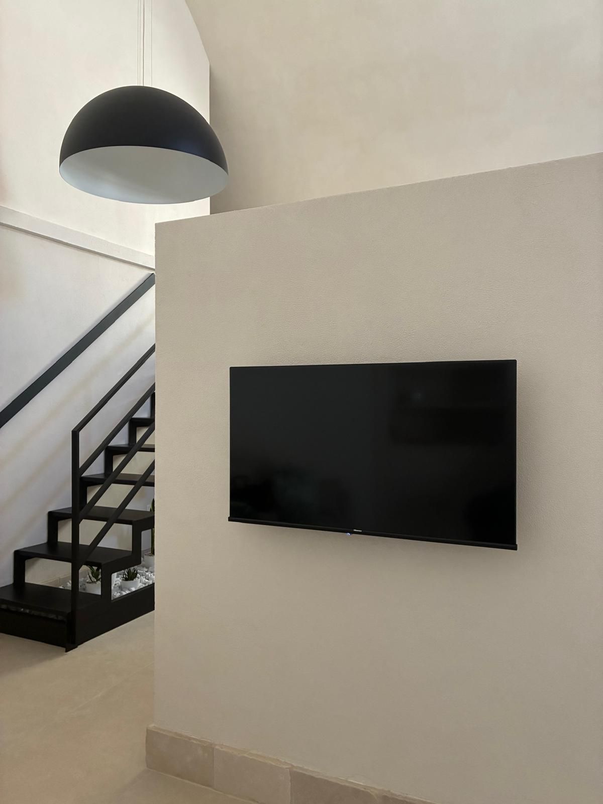 Ein modernes Interieur mit einem an der Wand montierten Flachbildfernseher, einer Treppe mit schwarzen Geländern und einer Pendelleuchte.