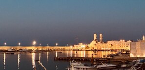 A waterfront marina with boats docked, a large illuminated building with a tower, and a calm sea at night.