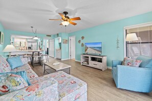 A living room with a ceiling fan, a television, a dining area with a table and chairs, and a kitchen in the background. - Heavenly Sunsets by the beach! (Myrtle Beach)