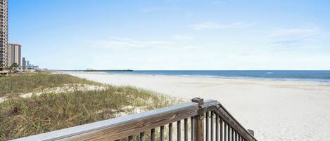 A wooden boardwalk leading to a sandy beach with a clear view of the ocean and distant buildings.