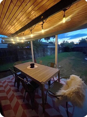 An outdoor patio with a wooden table and chairs, string lights, and a white fluffy object. - Cozy Home with Large Yard (Aurora)