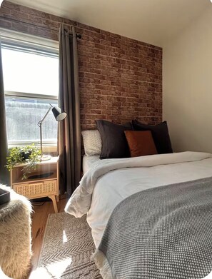 A bedroom with a brick wall, a bed with multiple pillows, a bedside table with a lamp, and a window with curtains. - Cozy Home with Large Yard (Aurora)