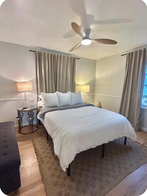 A bedroom with a bed, a bedside table, a lamp, a ceiling fan, and a window with curtains. - Cozy Home with Large Yard (Aurora)