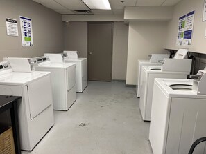 A laundry facility with multiple washing machines and dryers, a door, and informational posters on the walls. - Walk to Creekside Gondola – Cozy 1BR Condo w/ Patio, Pool & Hot Tub Access (Whistler)