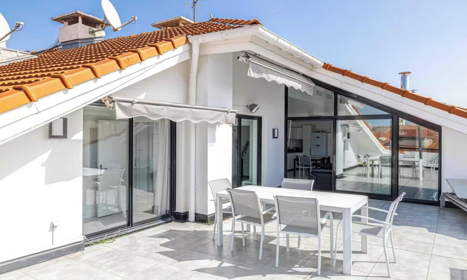 Una terraza moderna en la azotea con zona de comedor, puertas de cristal que dan a una cocina y tejado de baldosas.