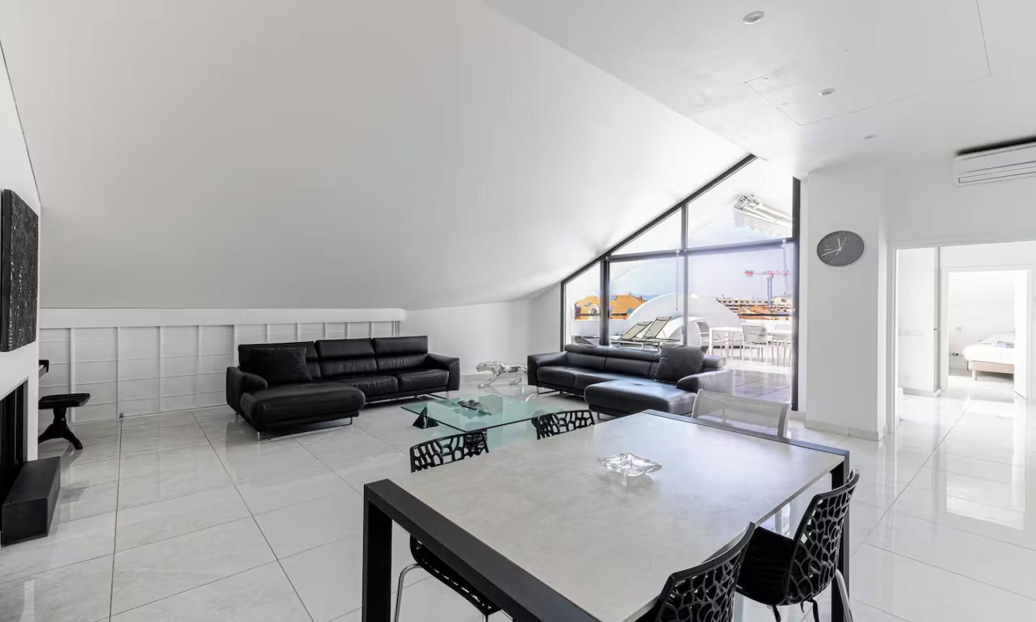 A modern living room with a black leather sofa, a glass coffee table, and a dining area with a white table and chairs.