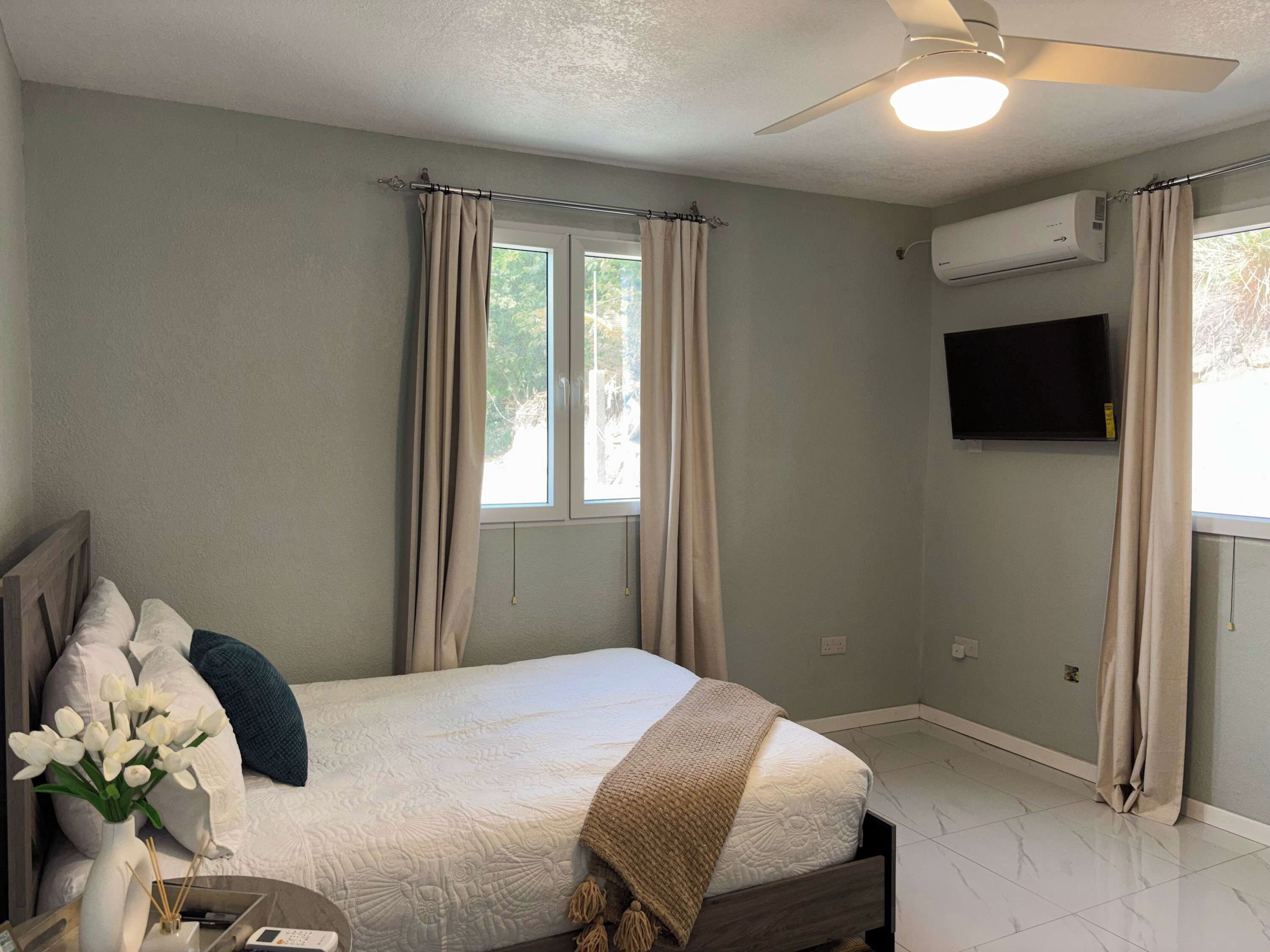 Une chambre à coucher comprenant un lit, des rideaux, un ventilateur de plafond, un appareil de climatisation, un téléviseur et une fenêtre.