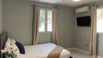Une chambre à coucher comprenant un lit, des rideaux, un ventilateur de plafond, un appareil de climatisation, un téléviseur et une fenêtre.