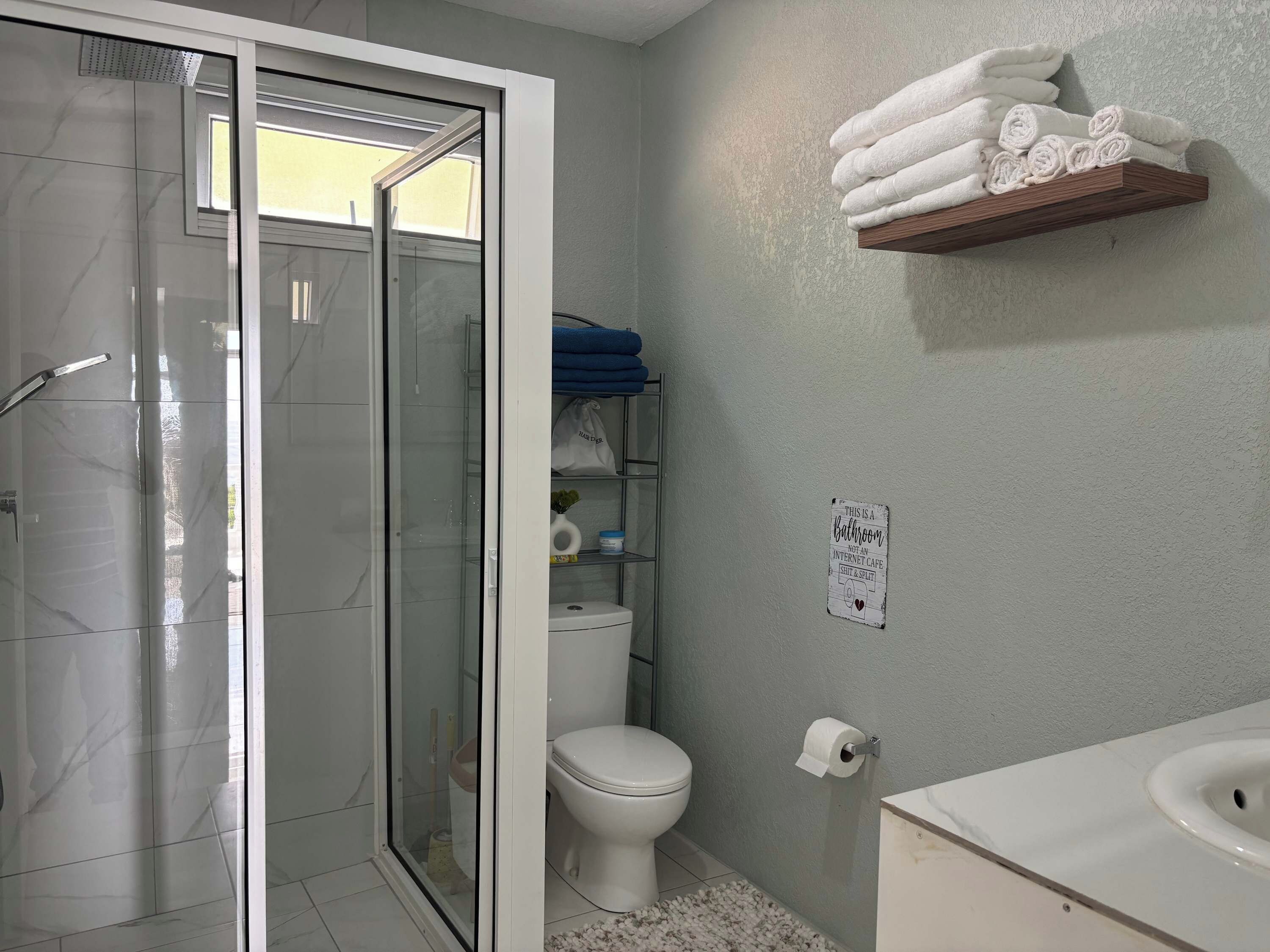 Une salle de bain avec une douche à parois de verre, des toilettes, une étagère avec des serviettes et un panneau sur le mur.