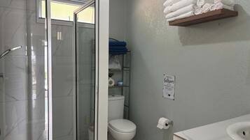 Une salle de bain avec une douche à parois de verre, des toilettes, une étagère avec des serviettes et un panneau sur le mur.