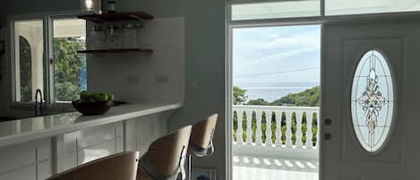 A modern kitchen with a large window, two bar stools, and a decorative door with a view of the ocean.