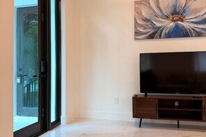 Habitación con una televisión de pantalla grande sobre un soporte de madera, una puerta corrediza de vidrio que da al exterior y un cuadro colgado en la pared.