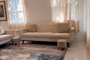 Una sala de estar con un sofá color beige, dos otomanes a juego, una alfombra estampada y cortinas translúcidas.