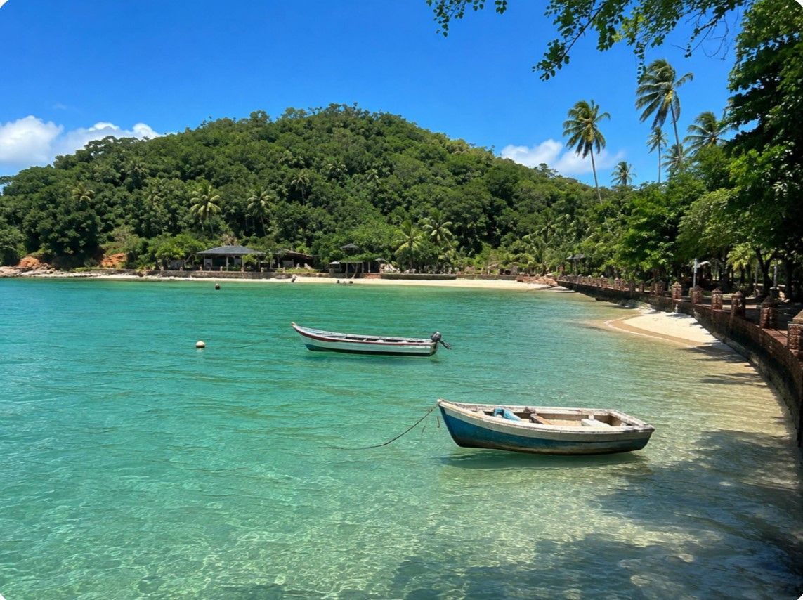 A beach with clear turquoise water, two boats anchored near the shore, and a backdrop of lush green hills.