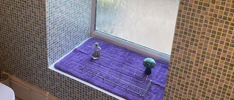 Casa de banho com janela, um porta-toalhas roxo e uma pequena prateleira com uma garrafa e um objeto decorativo.
