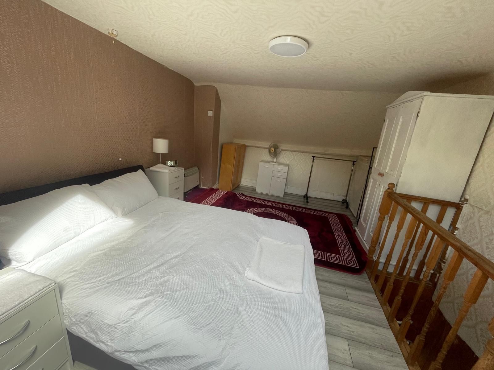 A bedroom with a bed, a nightstand, a dresser, a red patterned rug, a wooden railing, and a ceiling light.