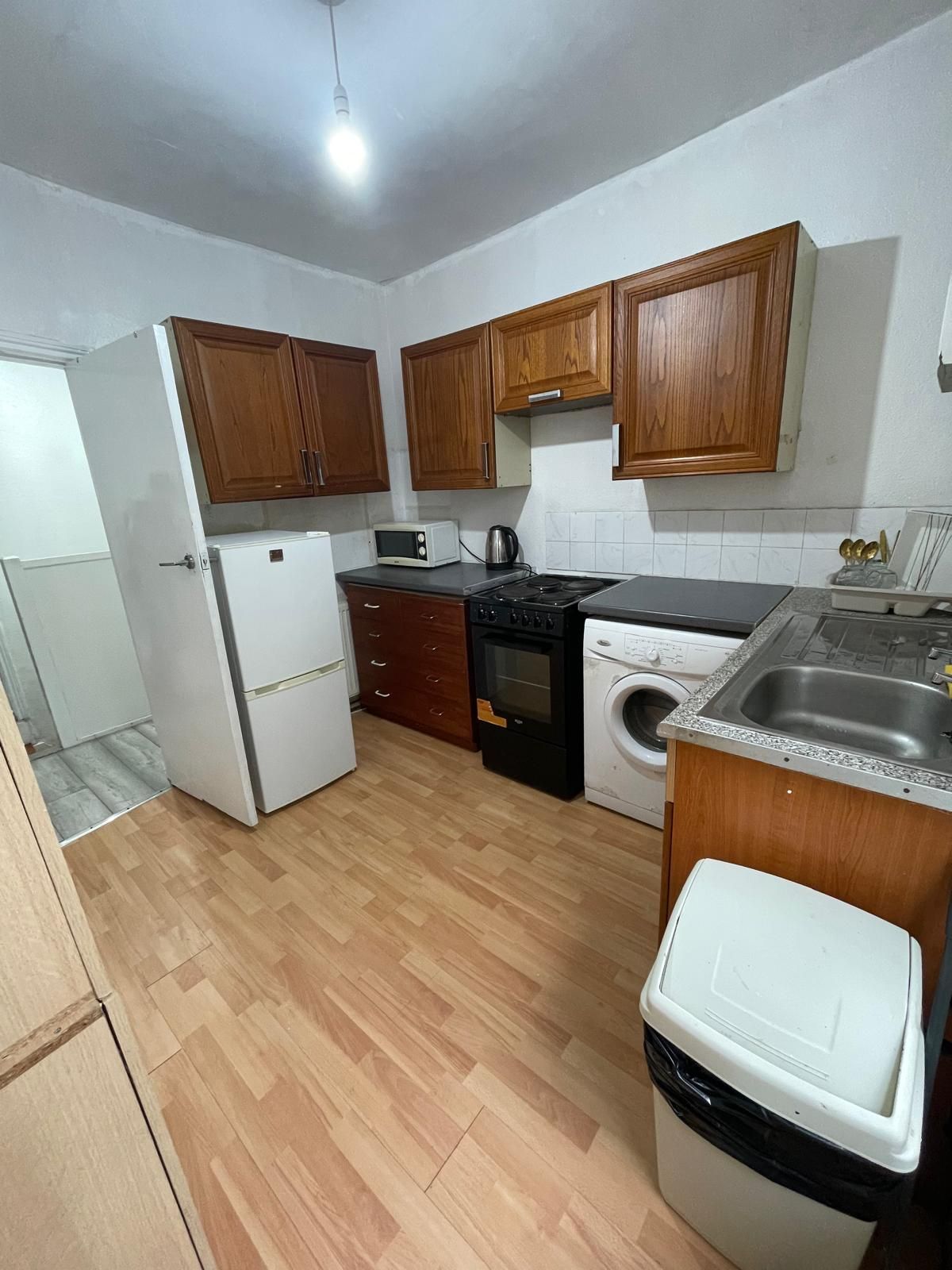A compact kitchen with wooden cabinets, a refrigerator, a microwave, a stove, a washing machine, and a sink.
