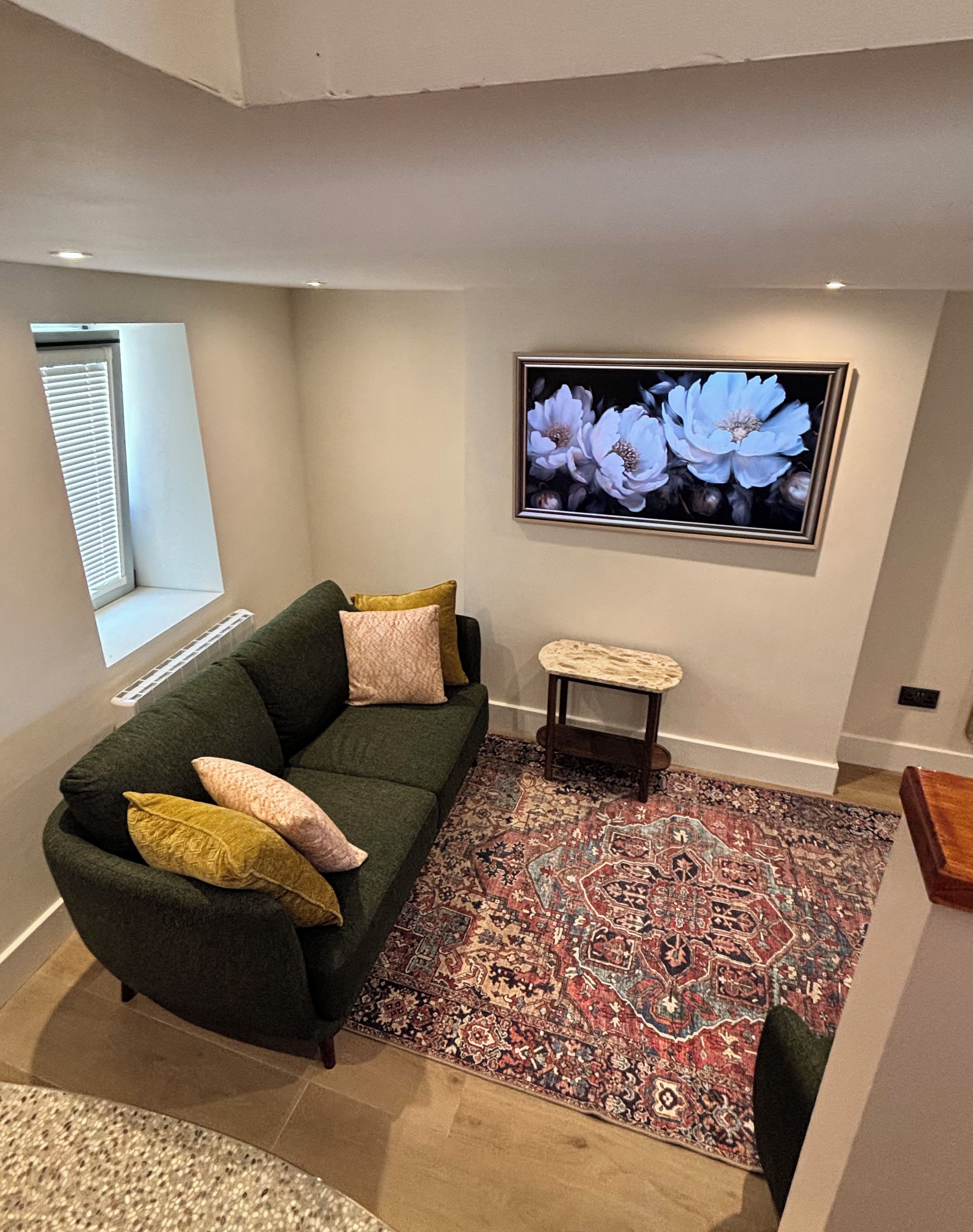 Un salon avec un canapé vert, un tapis à motifs et une œuvre d’art florale encadrée accrochée au mur.