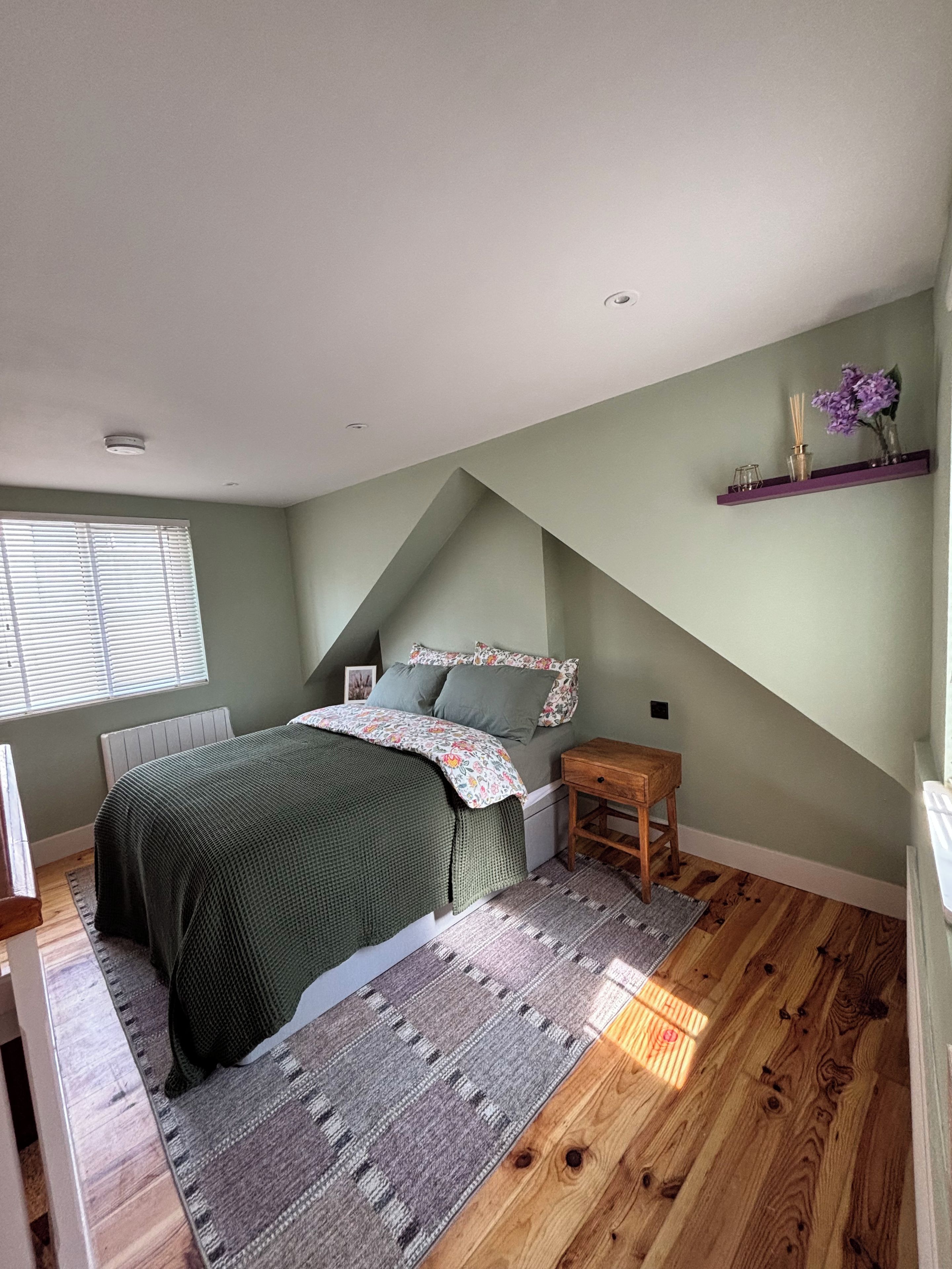 A bedroom with a sloped ceiling, a bed with a floral bedspread, a wooden nightstand, a small table, a window with blinds, and a purple shelf with decorative items.