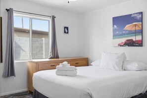 Ein Hotelzimmer mit einem Bett, einer Holzbank, einem Fenster mit Vorhängen und einem Gemälde mit Strandmotiv an der Wand.